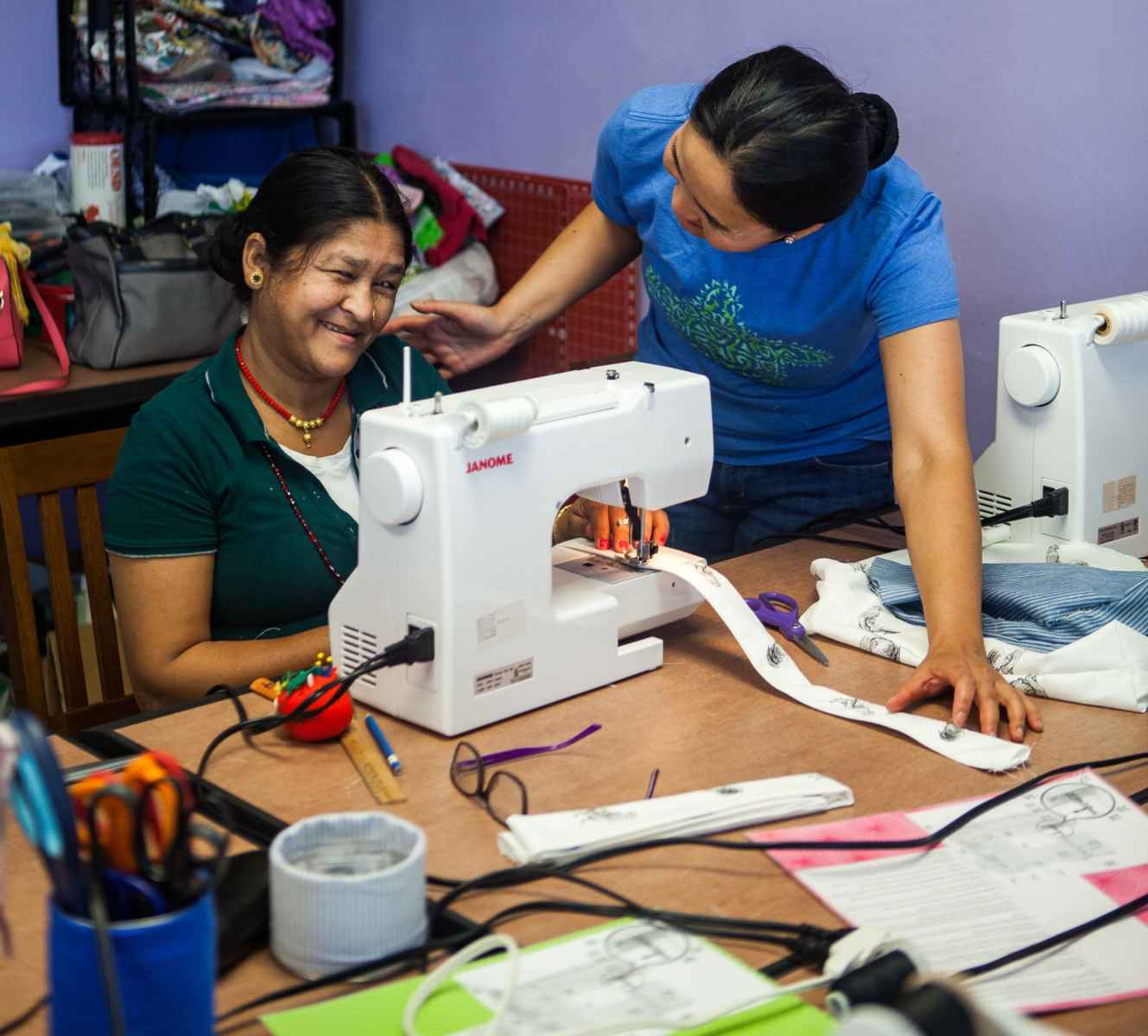 Smiling woman sewing with guidance from another woman holding the fabric